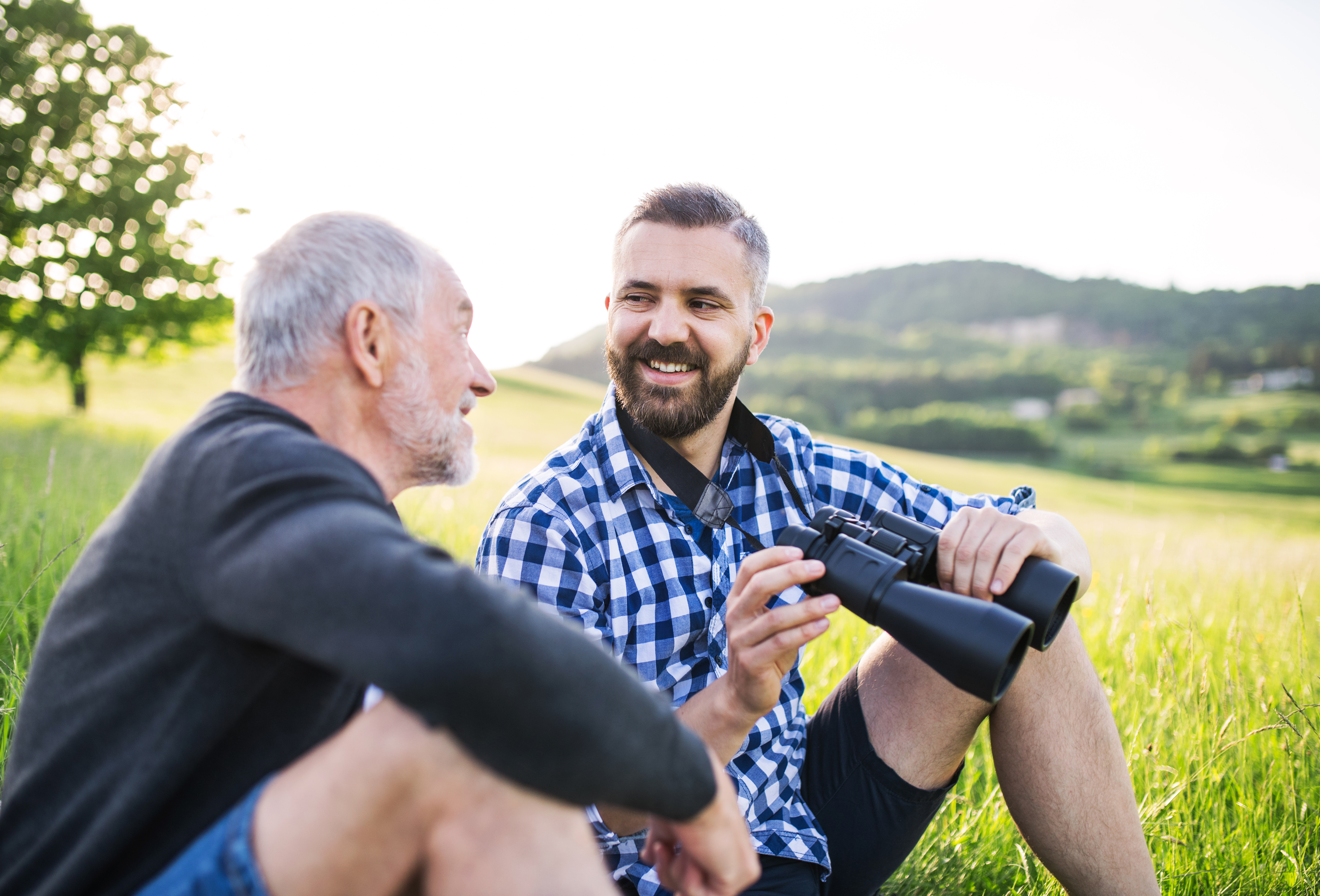 An adult hipster son with binoculars and senior father sitting on the grass in sunny nature.